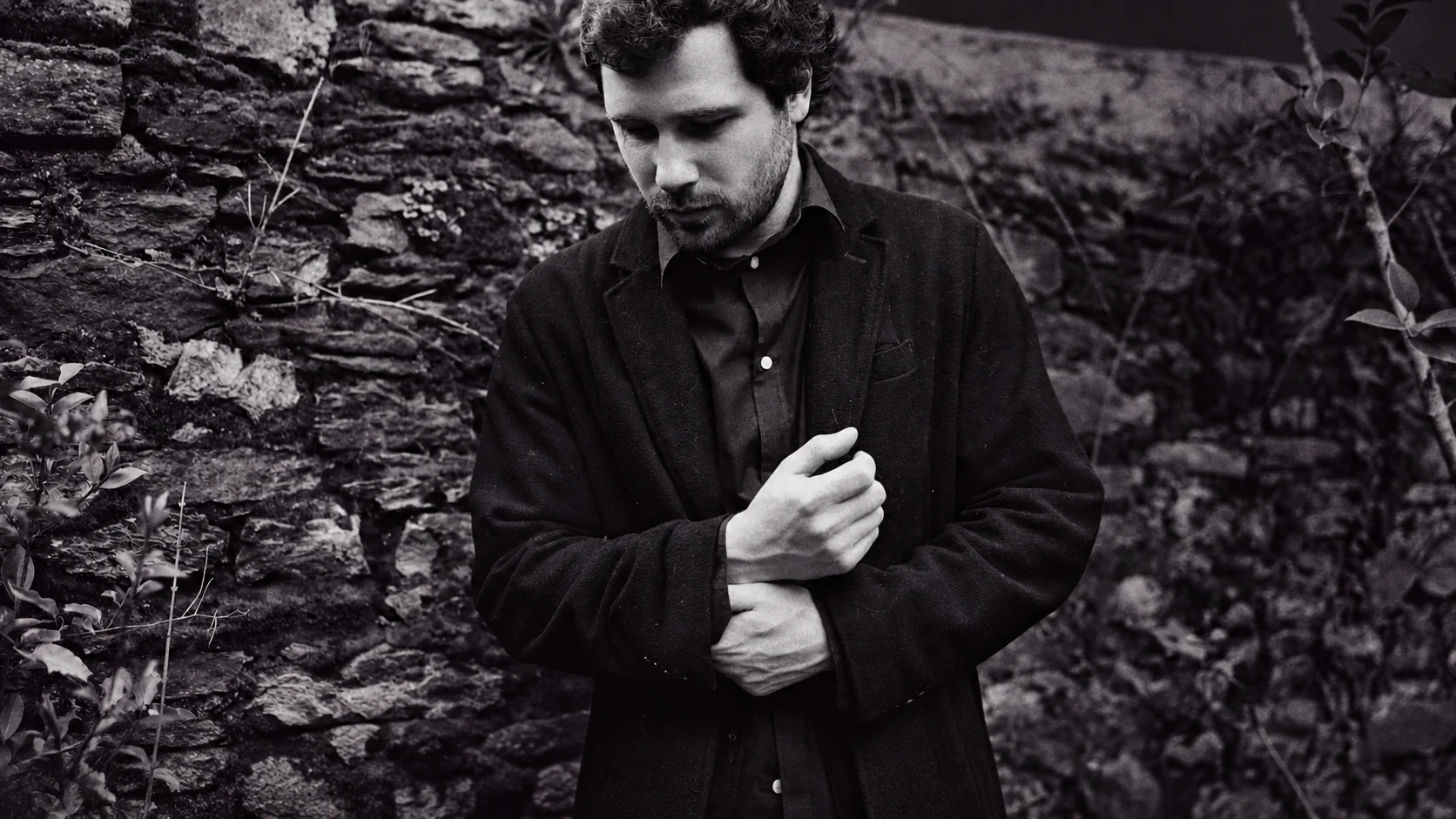 Black and white portrait of Giorgi Koberidze in a dark coat, head bowed, in front of an old stone wall